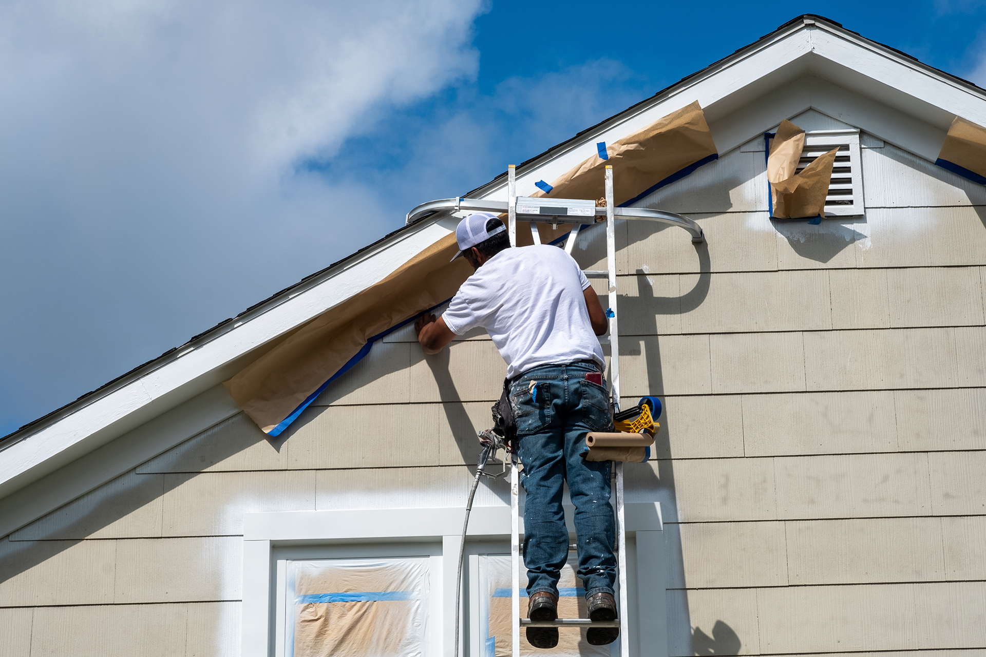 Painter using ladder and sprayer for two-story exterior house painting